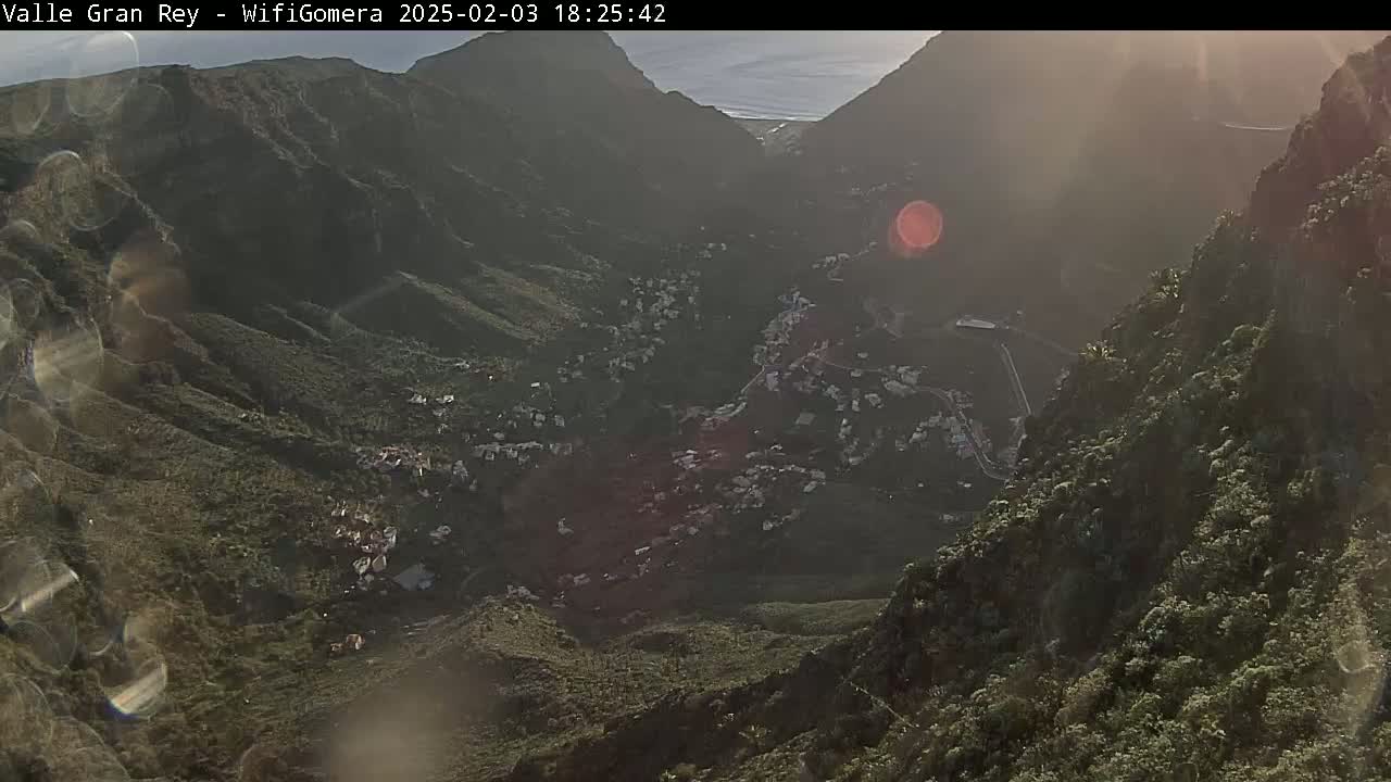 A sunlit village nestled in a valley between steep, verdant mountainsides.