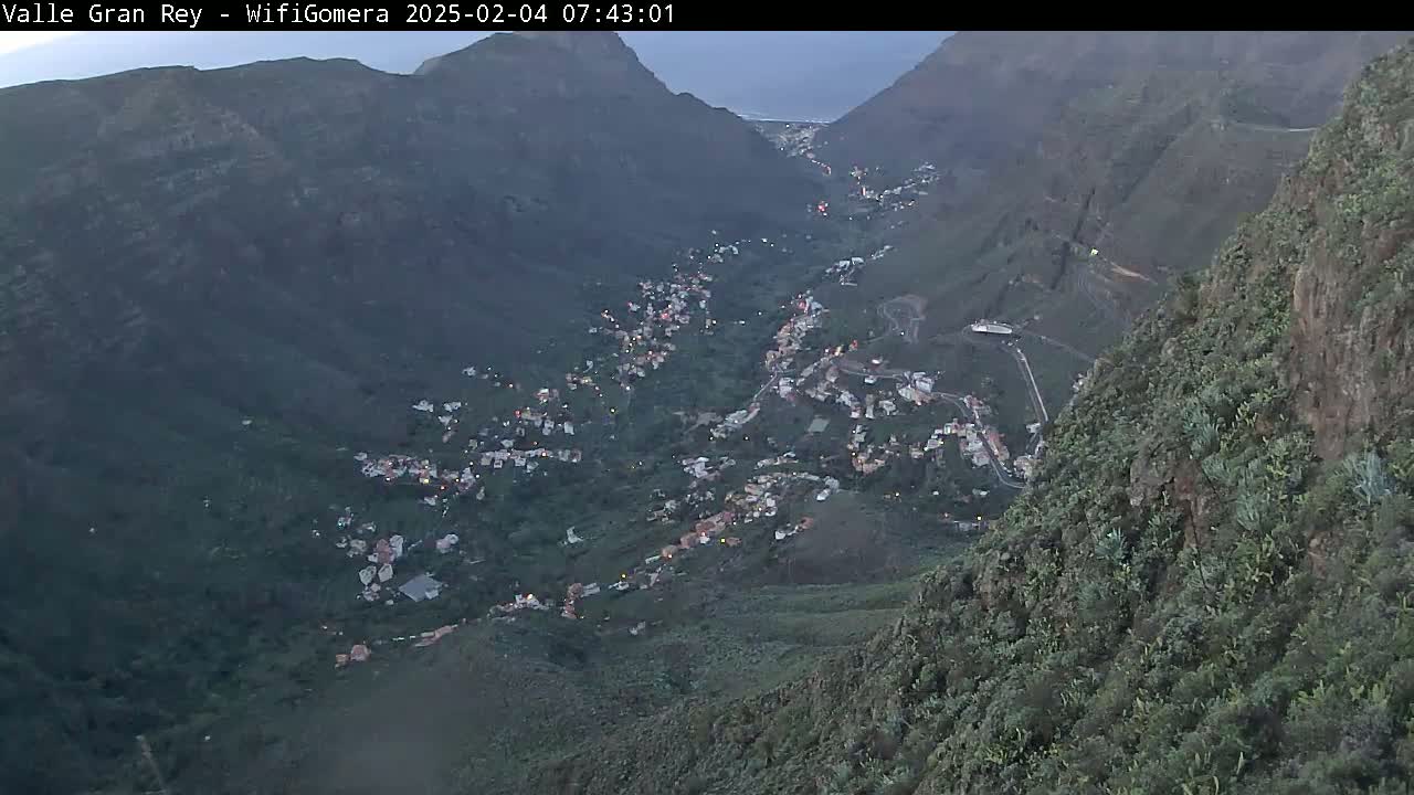 A mountain valley town at dusk is nestled between steep, verdant hillsides under a mostly clear sky.