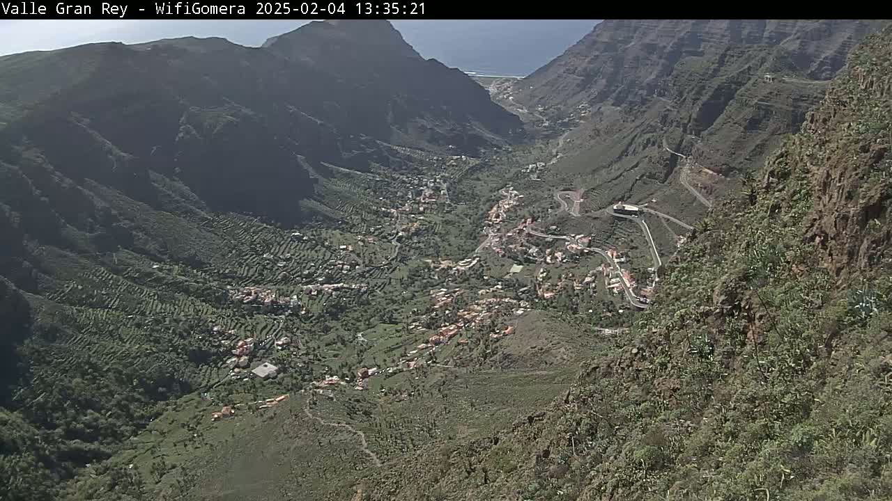 A high-angle view shows a valley town nestled between steep, rocky mountains on a sunny day.