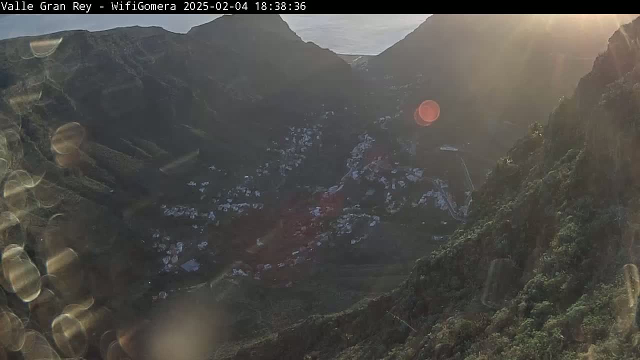 A high-angle, hazy view at sunset shows a village nestled in a valley between steep, dark green mountains.