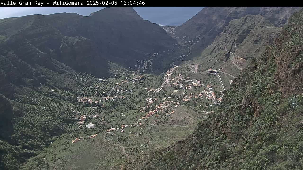 A high-angle view on a sunny day shows a village nestled in a verdant valley between steep, rocky mountainsides.