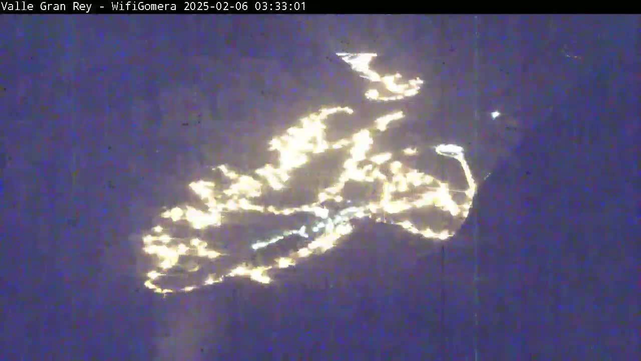 A nighttime, aerial view shows the lights of a town or city reflected on a dark body of water.