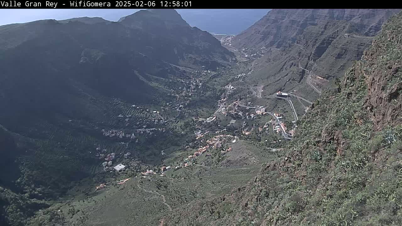A high-angle view shows a village nestled in a verdant valley between steep, rocky mountains under a mostly clear sky.