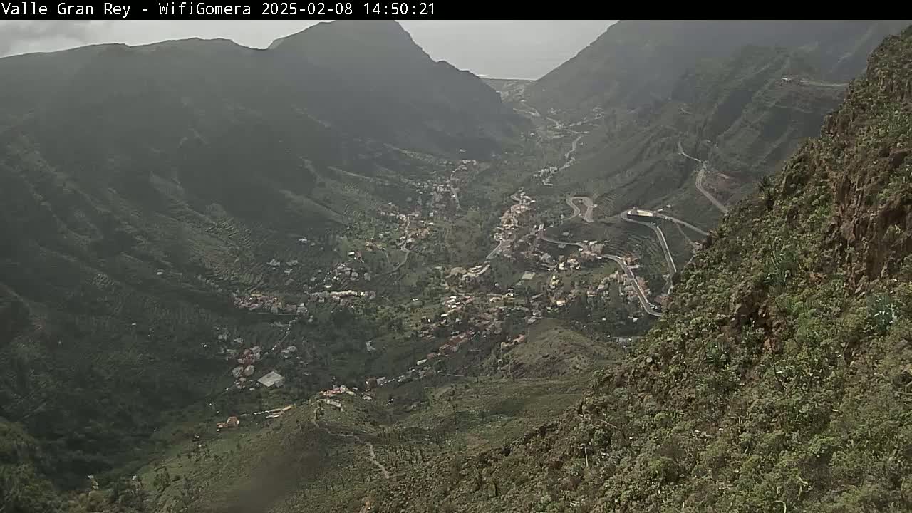 A hazy view from a mountaintop shows a valley with a town nestled among green hills and winding roads.