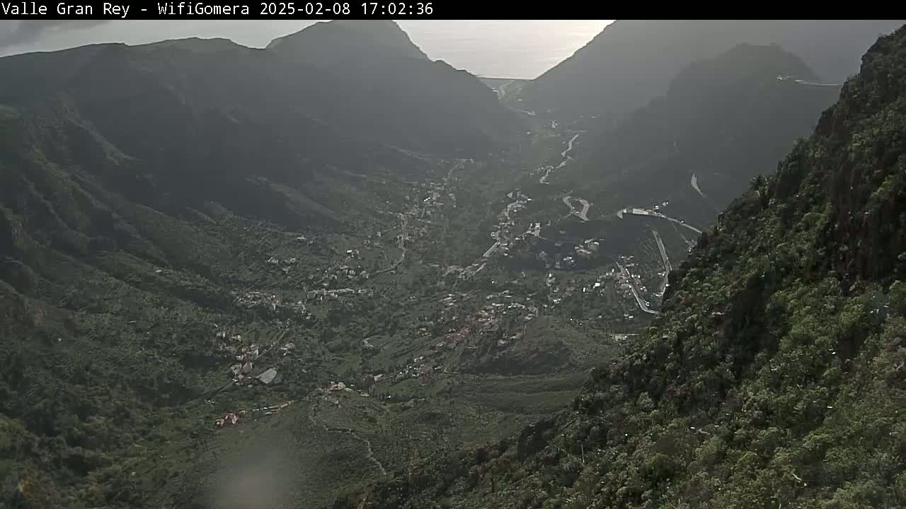 A hazy, high-angle view shows a valley town nestled between steep, verdant mountainsides with a winding road visible.