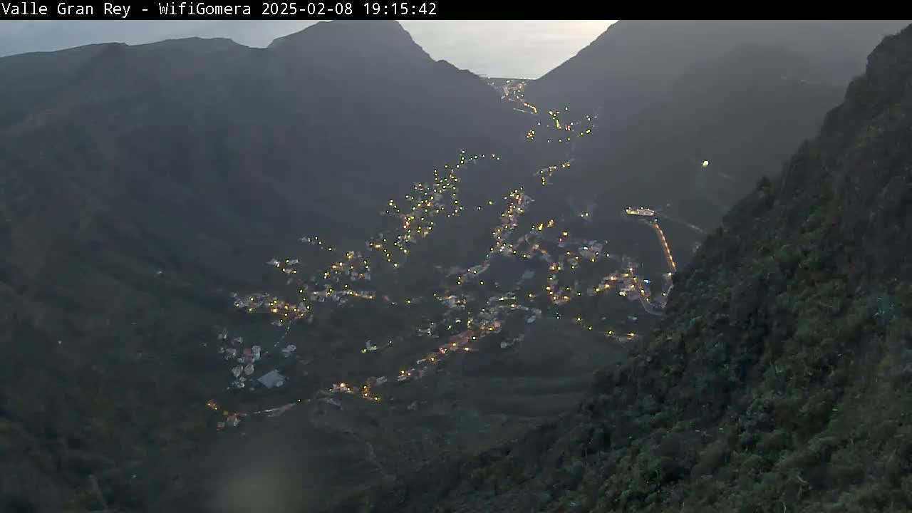 A valley nestled between dark mountains at night shows a town illuminated by scattered lights along a winding road.