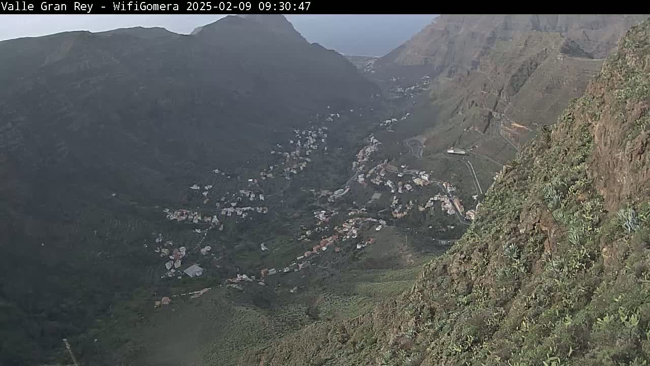 A hazy view from a mountaintop reveals a valley town nestled among green hills and rocky slopes.