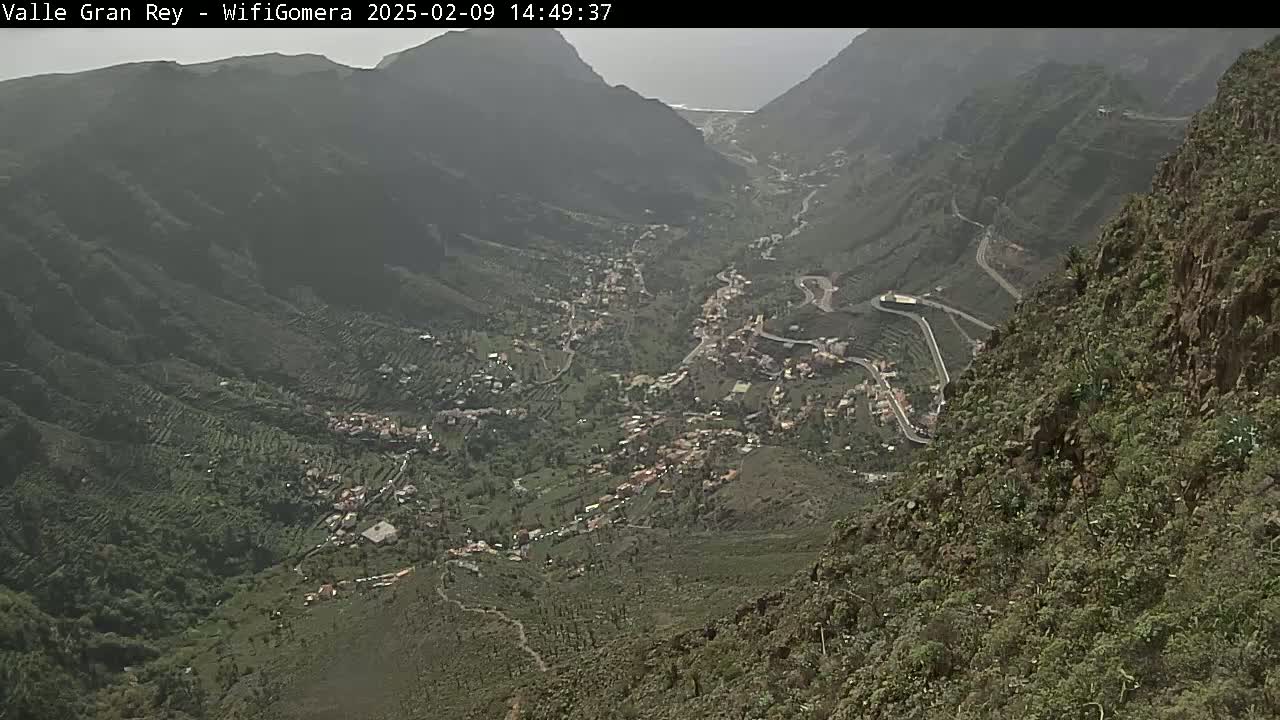 A hazy view from a mountaintop shows a valley with a town nestled among terraced hillsides and winding roads.