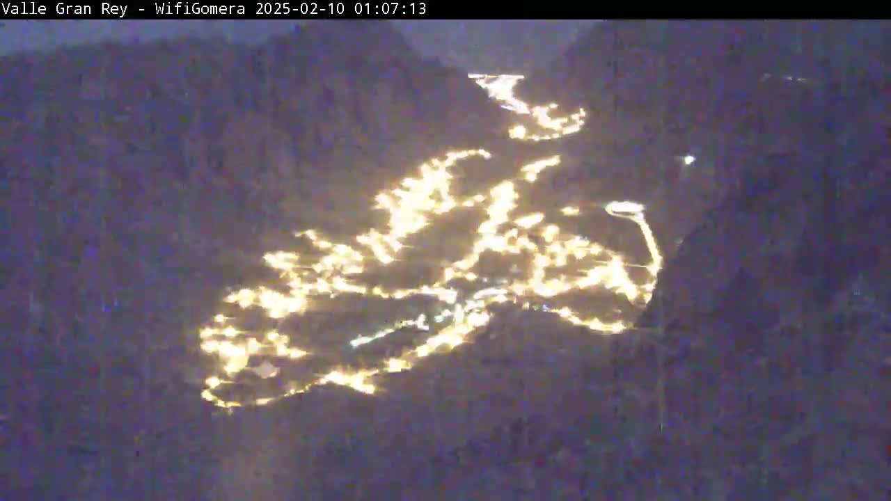 A nighttime, aerial view shows a valley illuminated by scattered lights winding along a road.