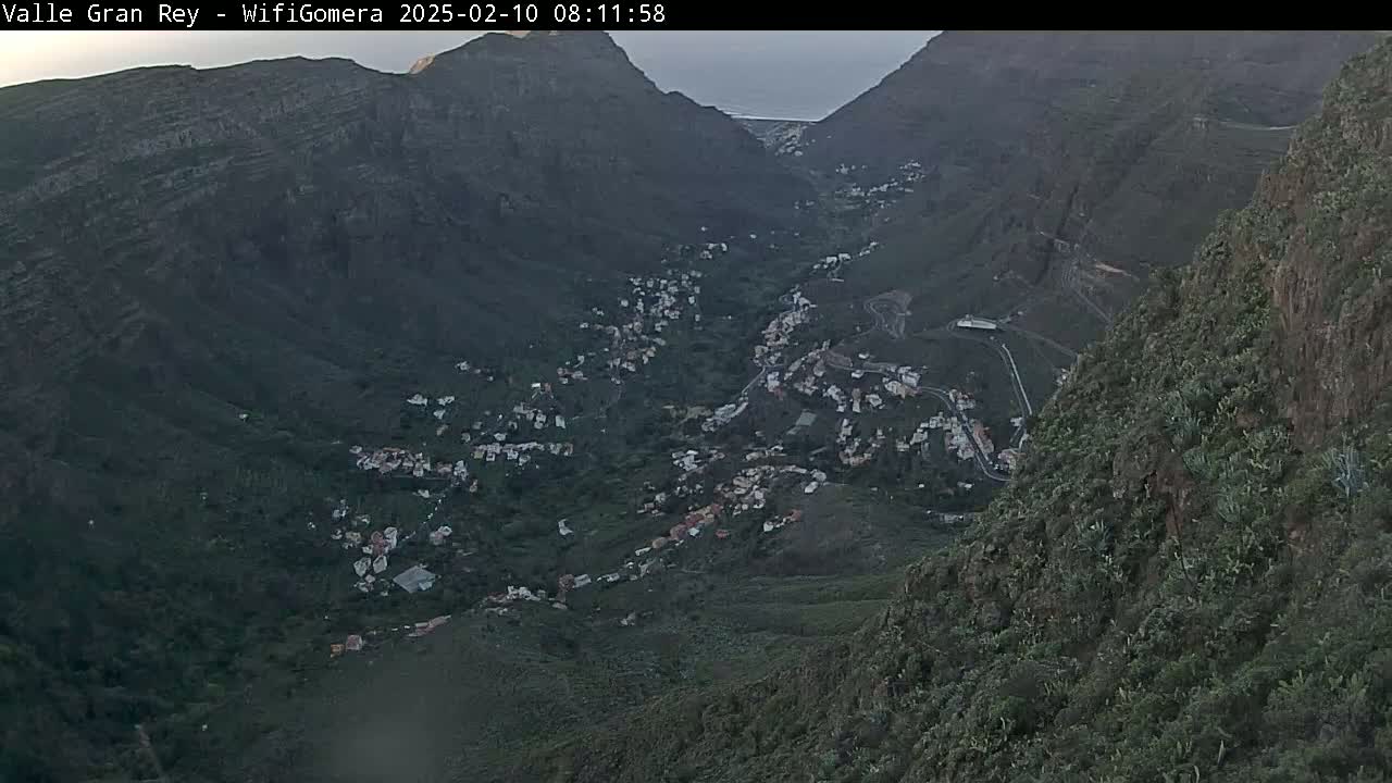 A high-angle view shows a valley town nestled between dark, steep mountainsides under a dusky sky.