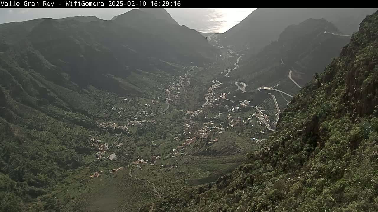 A high-angle, long shot shows a town nestled in a valley between steep, green mountains on a partly cloudy day.