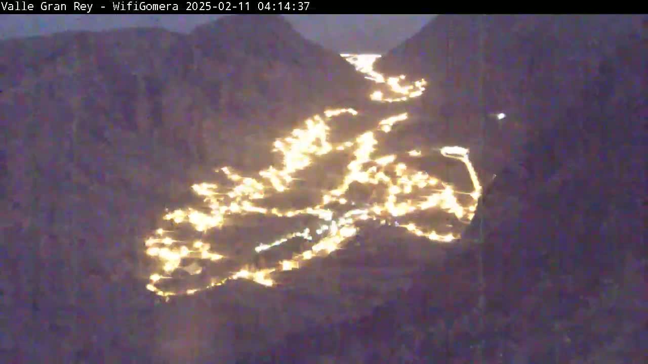 A nighttime aerial view shows a valley town's lights winding along a road between dark mountains.