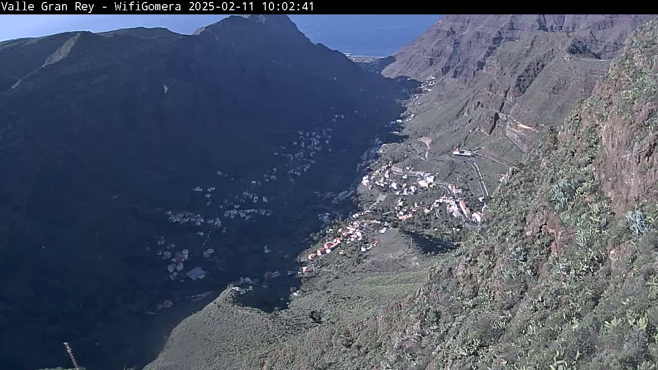 A sunny, high-angle view shows a valley nestled between steep, rocky mountains, with a small village situated in the valley bottom.