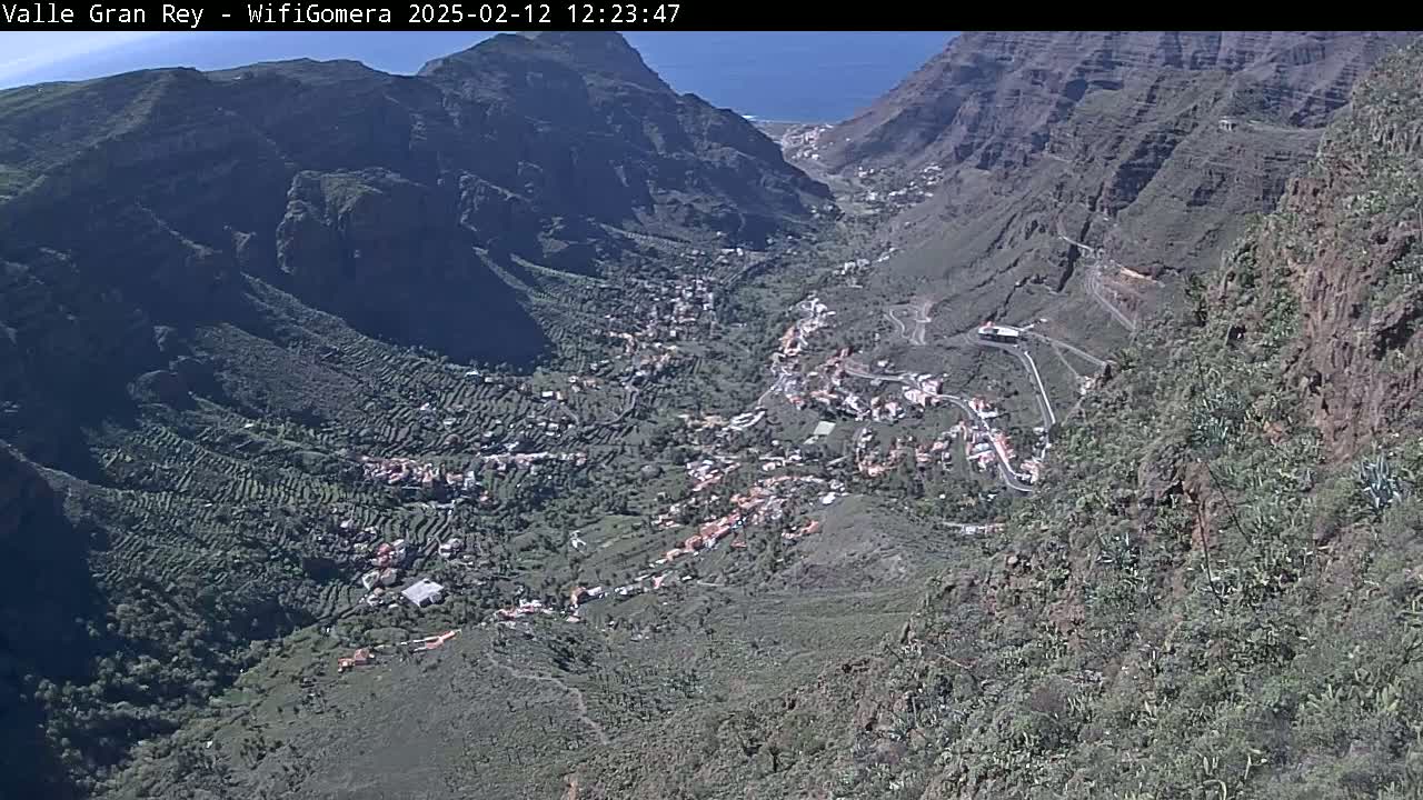 A high-angle view shows a village nestled in a mountainous valley on a sunny day, with the ocean visible in the distance.