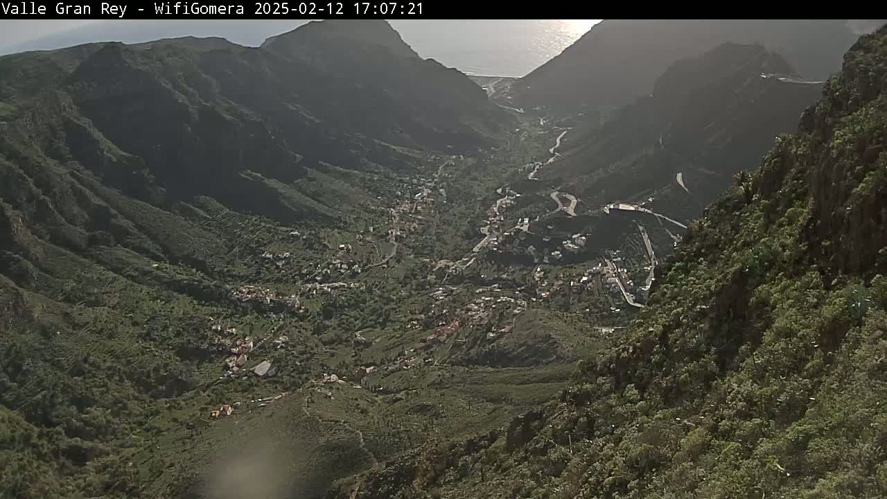 A high-angle, long shot shows a sunlit valley town nestled between steep, green-covered mountainsides, under a mostly clear sky.