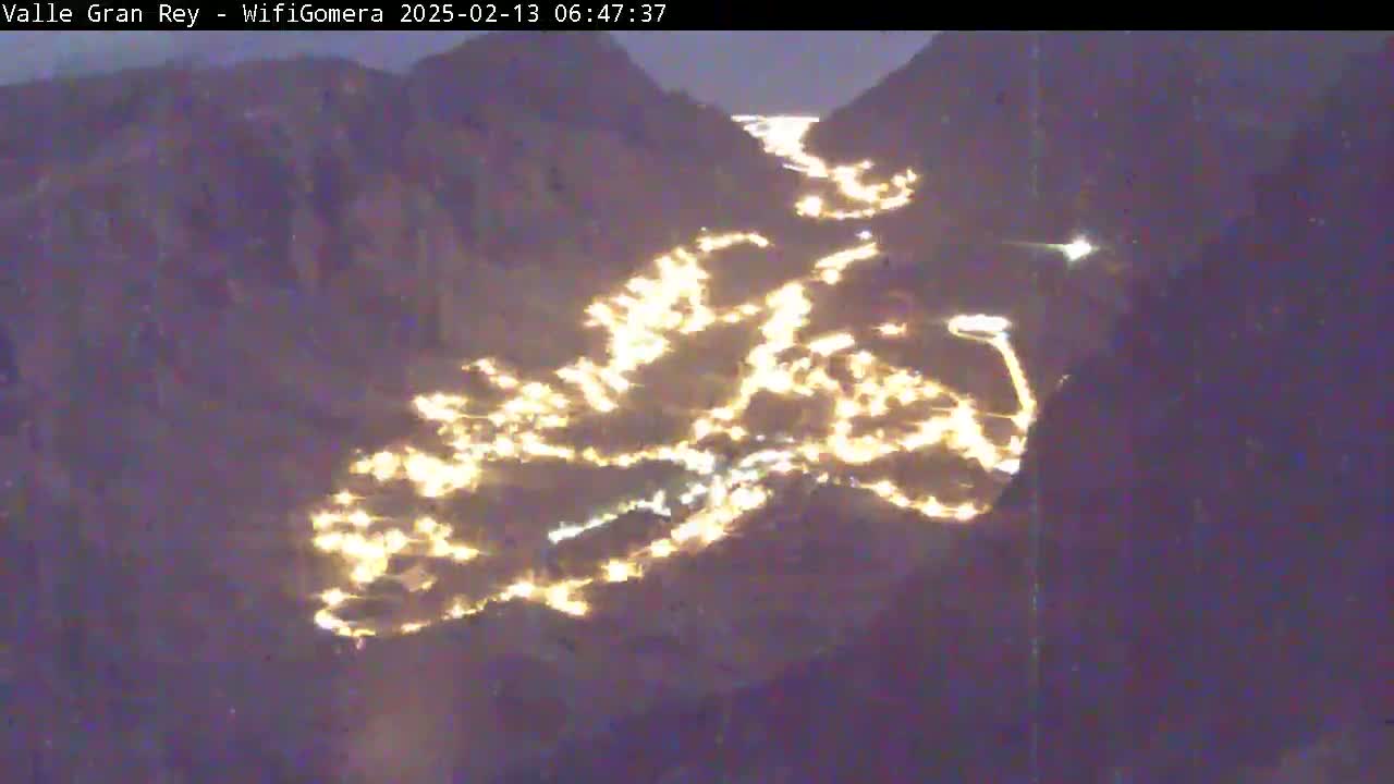 A nighttime, aerial view shows a town nestled in a valley between dark mountains, illuminated by numerous lights.