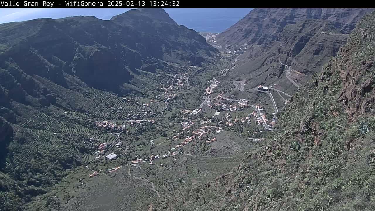 A sunny, high-angle view shows a village nestled in a steep, rocky valley, with winding roads snaking through the landscape.