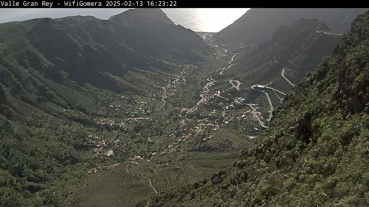A high-angle, long shot shows a village nestled in a verdant valley between steep, rocky mountains under a mostly clear sky, with a glimpse of the ocean in the far distance.