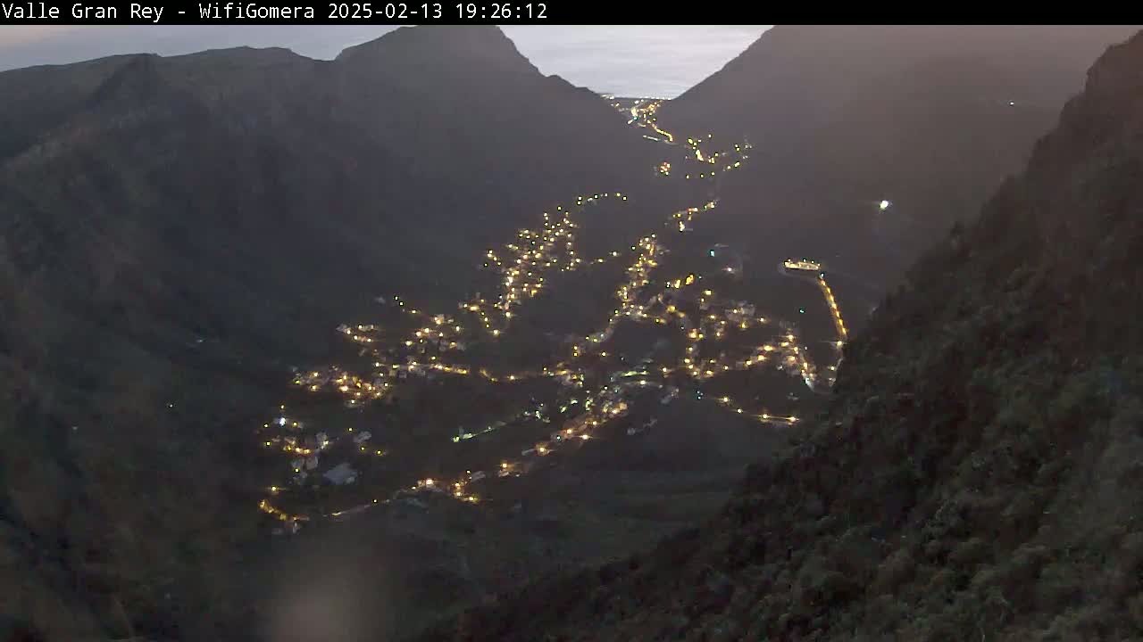 A nighttime, high-angle view shows a village nestled in a valley between dark mountains, illuminated by many small lights.