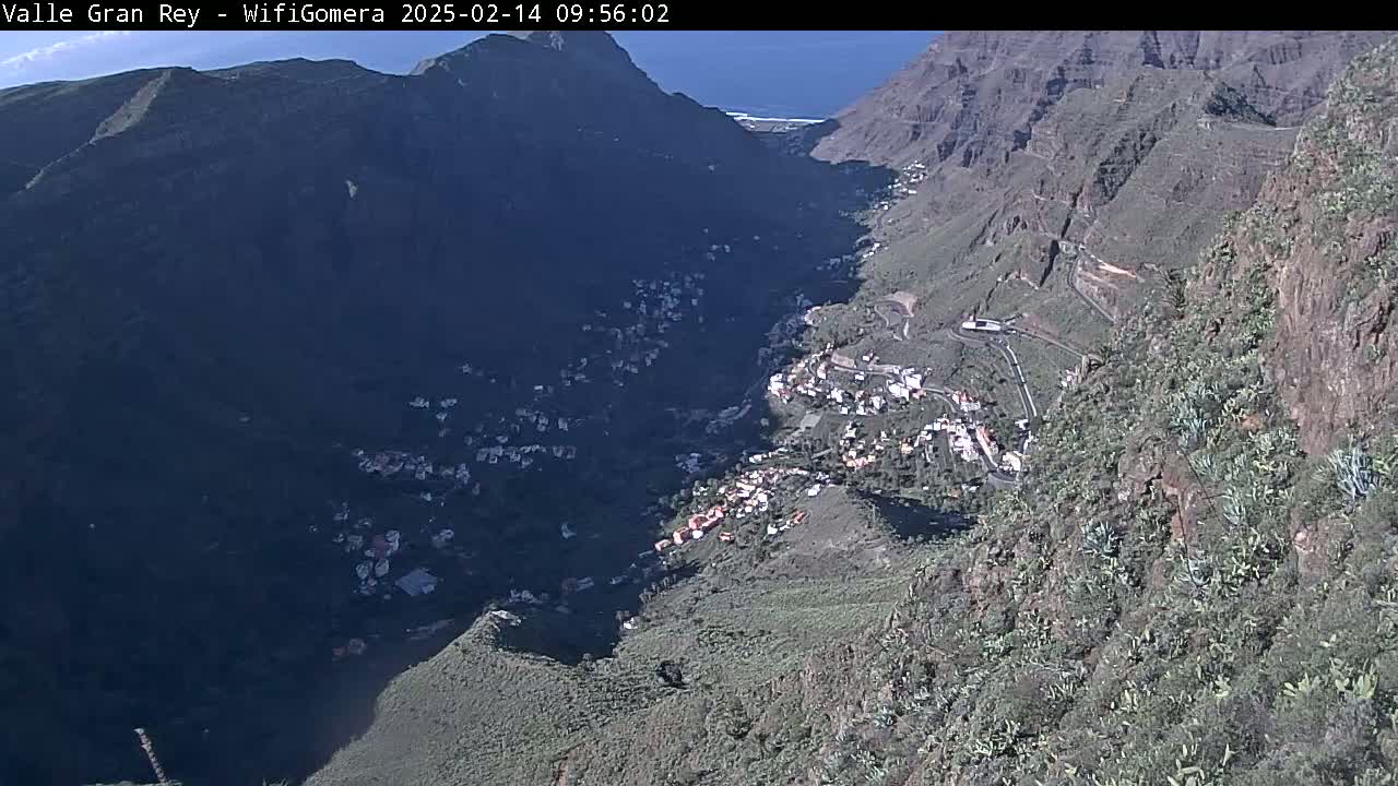 A high-angle, sunny view shows a mountainous valley with a small village nestled within, and a glimpse of the ocean in the distance.