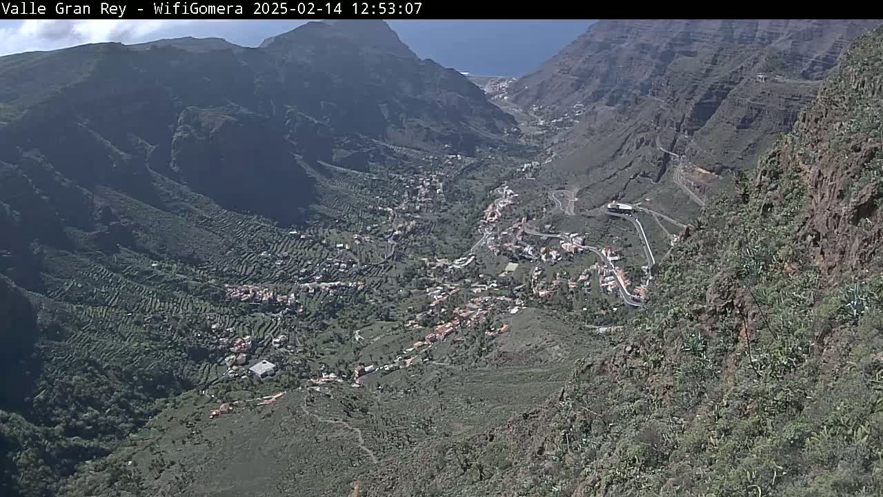A high-angle view shows a town nestled in a valley between steep, rocky mountains on a sunny day.