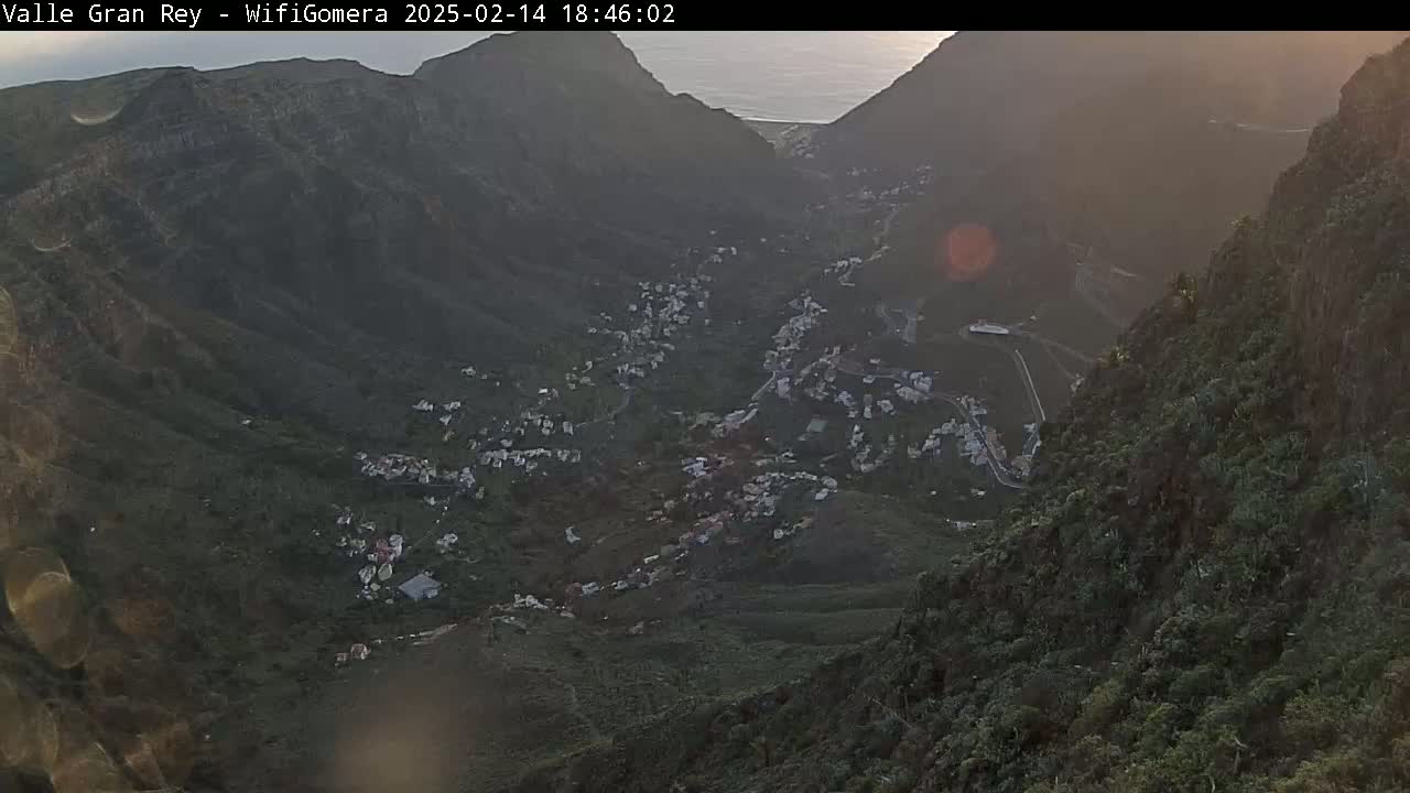 A high-angle view at sunset shows a village nestled in a valley between steep, dark green mountains, with a body of water visible in the distance.