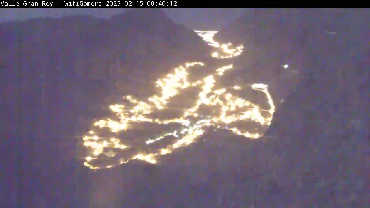 A nighttime aerial view shows a valley illuminated by scattered lights winding through a dark, mountainous landscape.