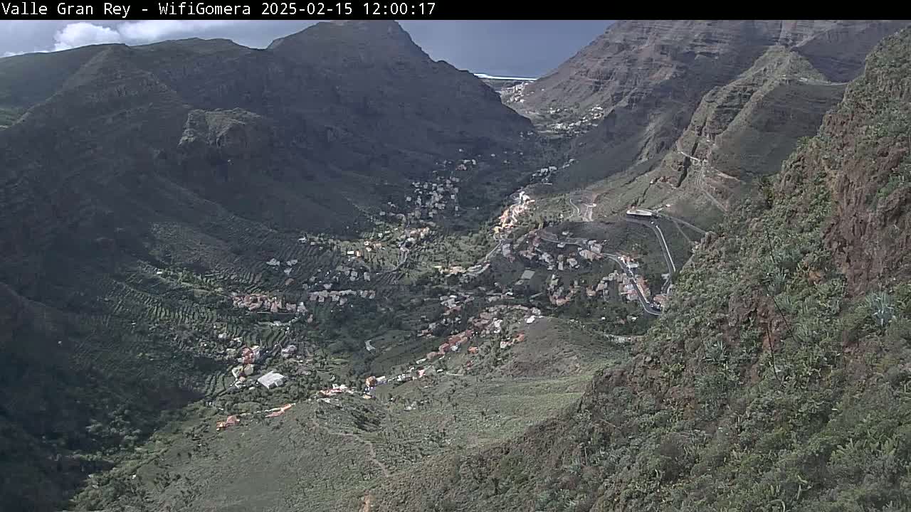 A high-angle view shows a valley nestled between steep, rocky mountains under a mostly sunny sky, with a small village and terraced farmland visible.