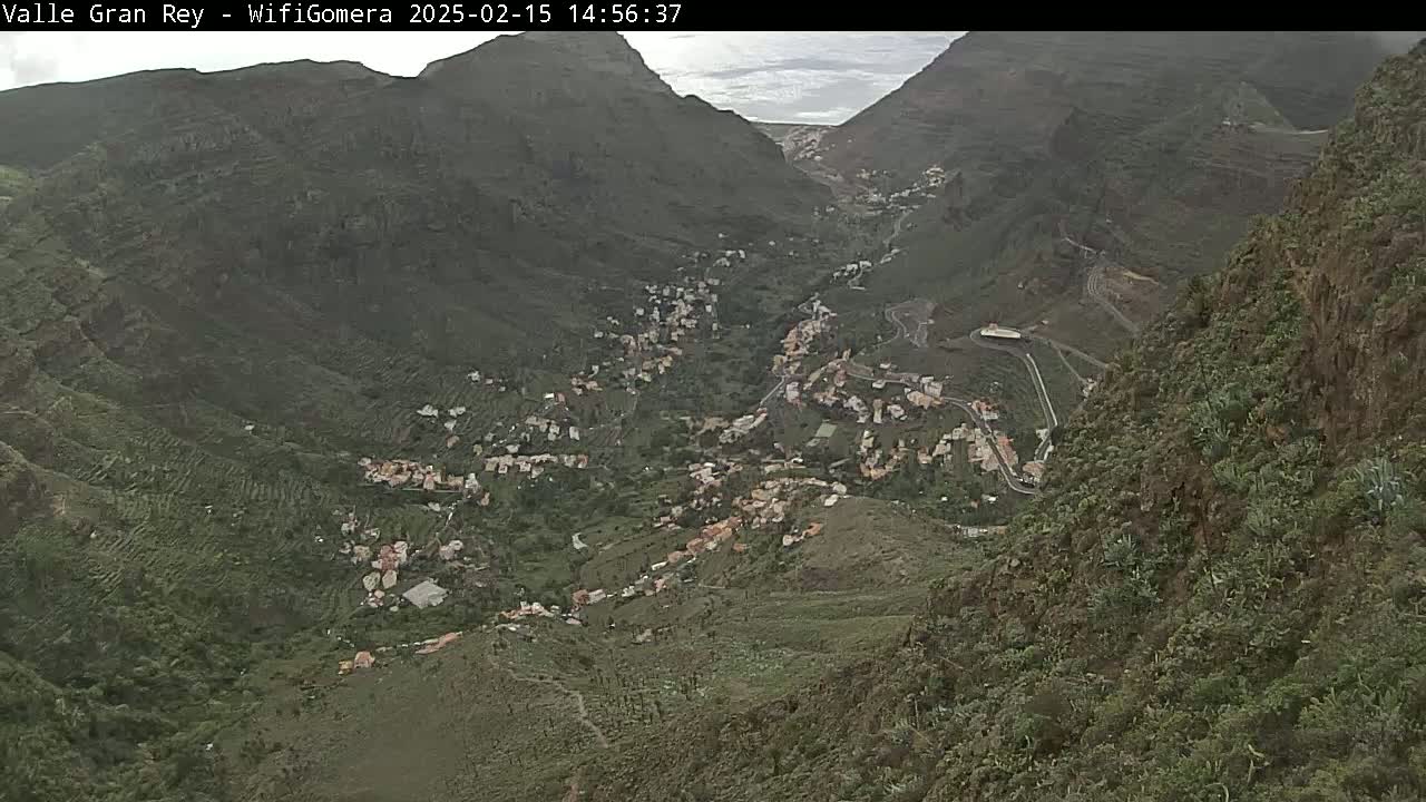 A high-angle view shows a town nestled in a verdant valley between steep, dark mountains under an overcast sky.
