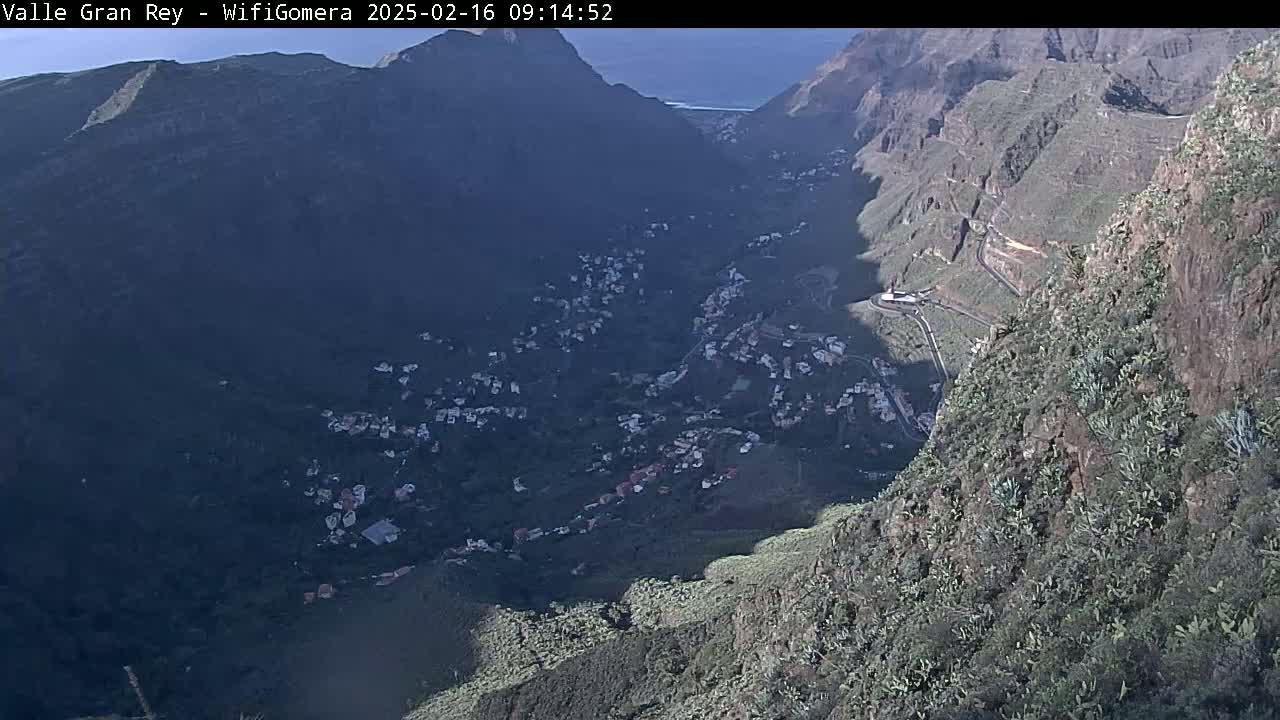 A high-angle view shows a valley nestled between steep, rocky mountainsides, with a small town spread across the valley floor under a partly sunny sky.