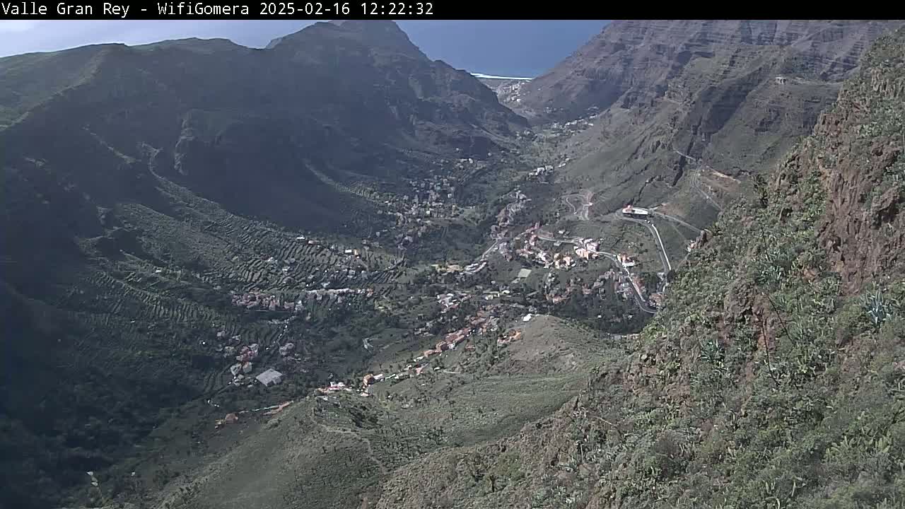 A high-angle view shows a valley town nestled between steep, rocky mountains under a clear, sunny sky.