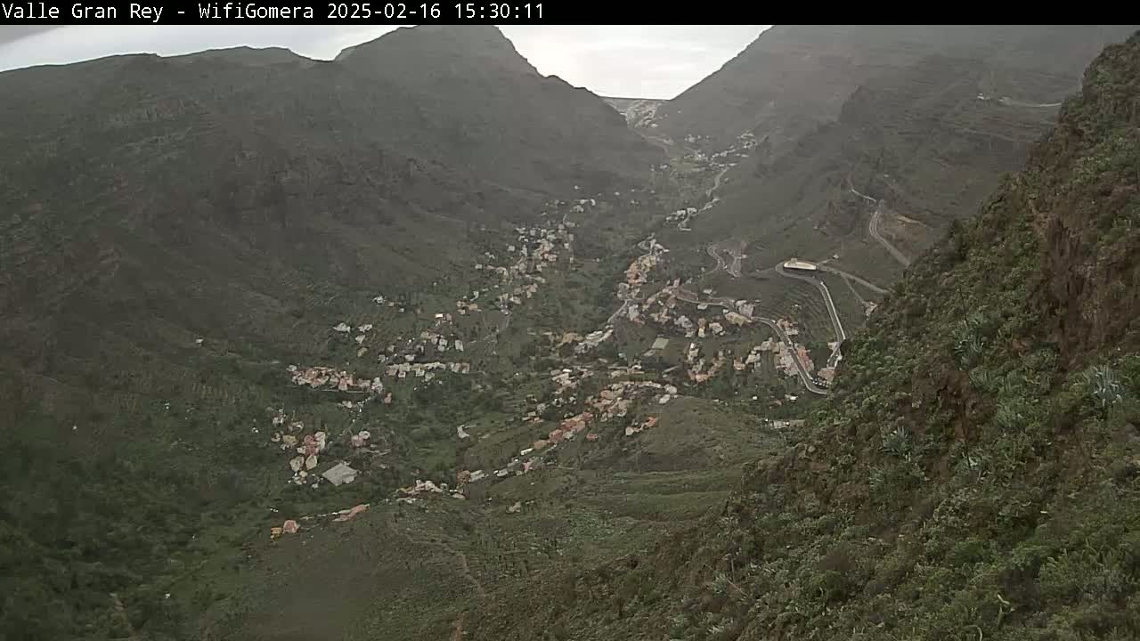 A mountainous valley town is seen on an overcast day, with winding roads snaking through the green and brown landscape.