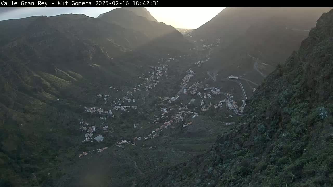 A high-angle, long shot shows a valley town nestled between steep, dark green mountains at dusk.