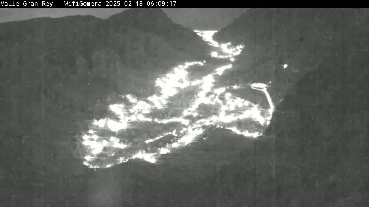 A nighttime, grayscale image shows a valley illuminated by bright, diffuse light sources appearing to be fires, nestled between dark mountains.