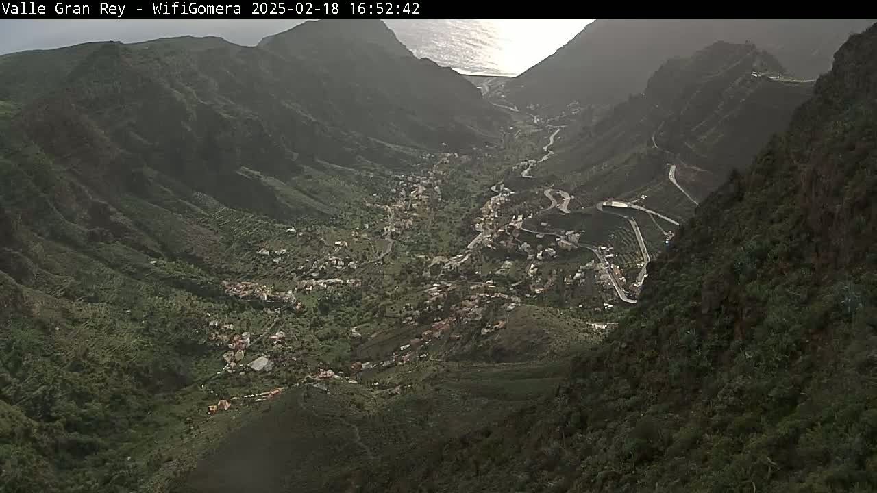 A high-angle view shows a verdant valley nestled between steep, dark mountainsides, partially cloudy with the ocean visible in the distance.