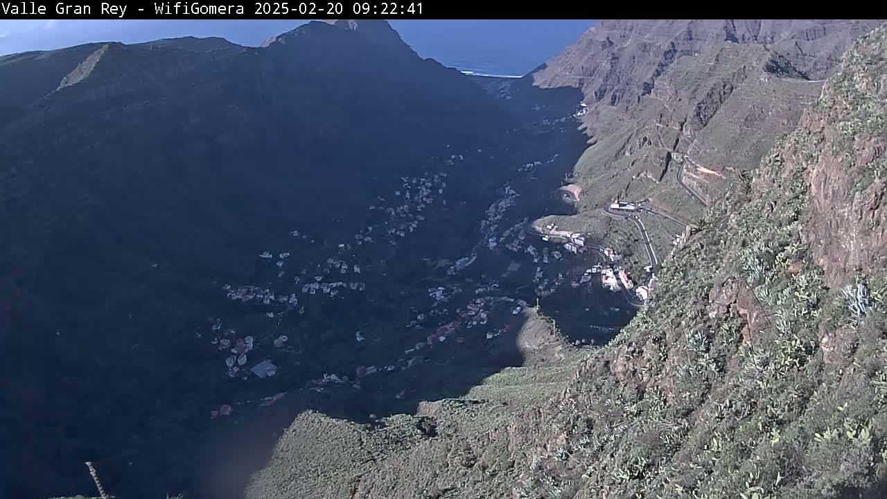 A high-angle view on a sunny day shows a small village nestled in a deep valley between steep, rocky mountainsides with sparse vegetation.