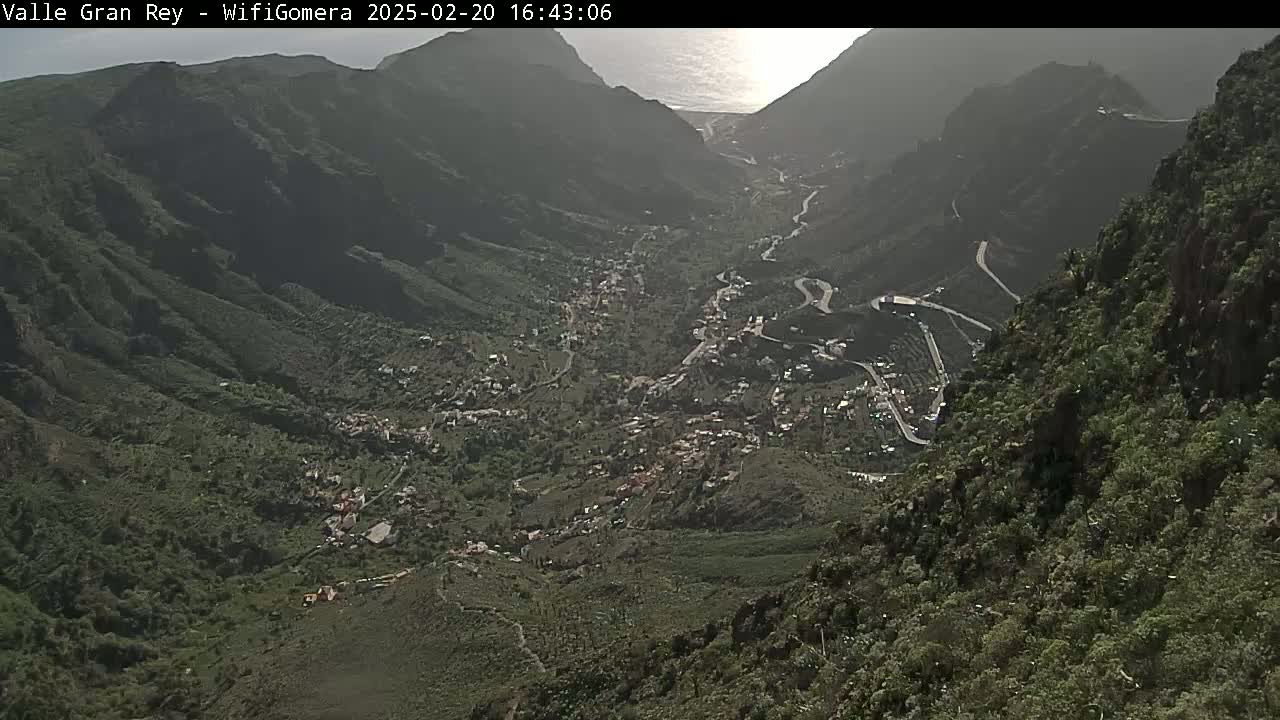 A high-angle, long shot shows a sunlit valley town nestled between steep, verdant mountainsides, with a winding road snaking through it and partially visible ocean in the distance.