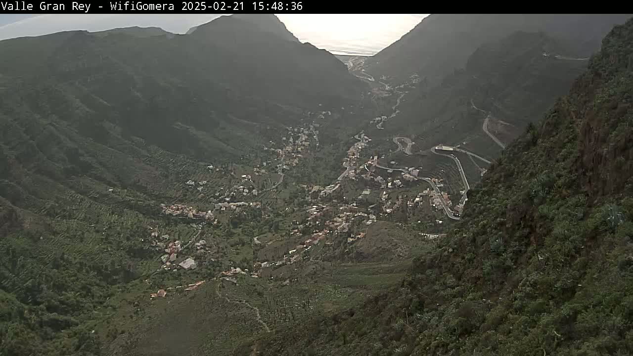 A hazy view shows a town nestled in a verdant valley between steep, dark mountains.