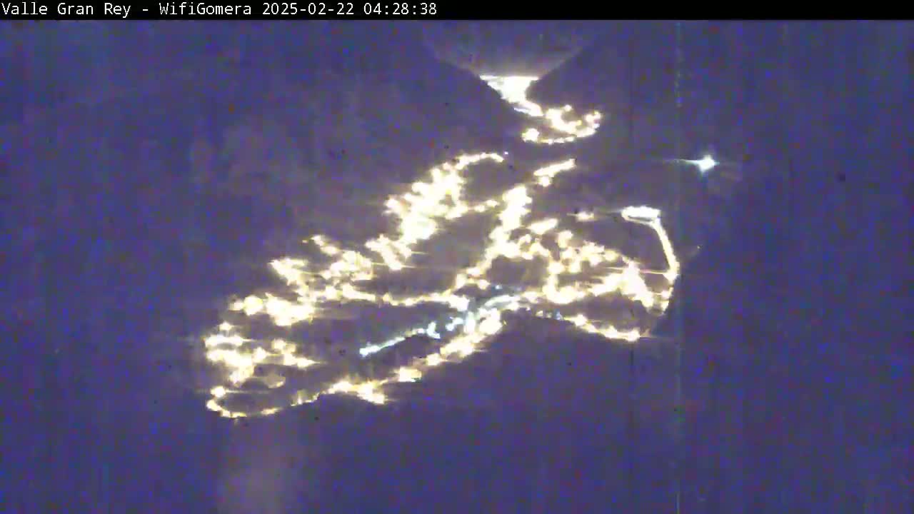 A nighttime, aerial view shows a valley illuminated by scattered lights, appearing to follow the contours of a winding road or river.