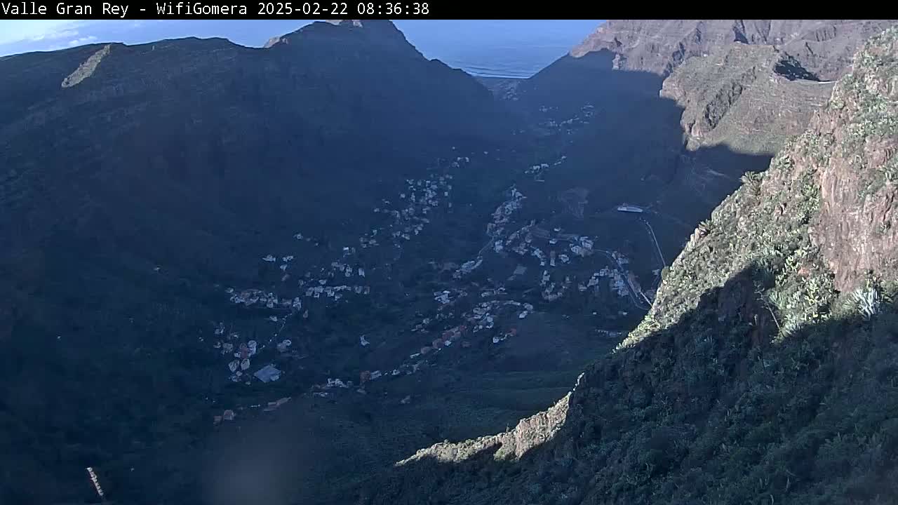 A high-angle view shows a valley town nestled between steep, dark mountains, partially illuminated by sunlight, with the ocean visible in the distance under a clear sky.