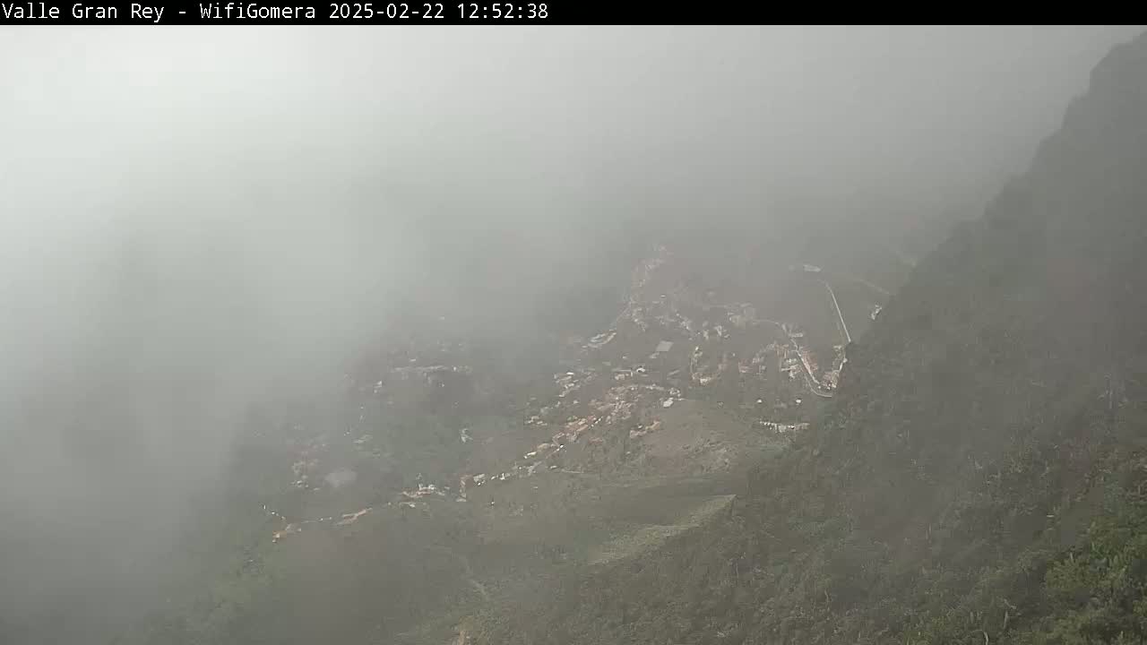 A foggy view shows a mountainside village nestled in a valley between steep, dark green hills.