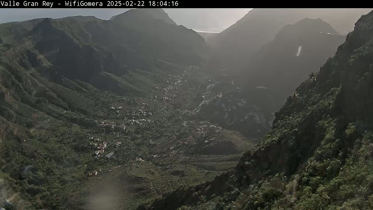 A hazy view shows a valley nestled between steep, green mountains under a mostly clear sky.