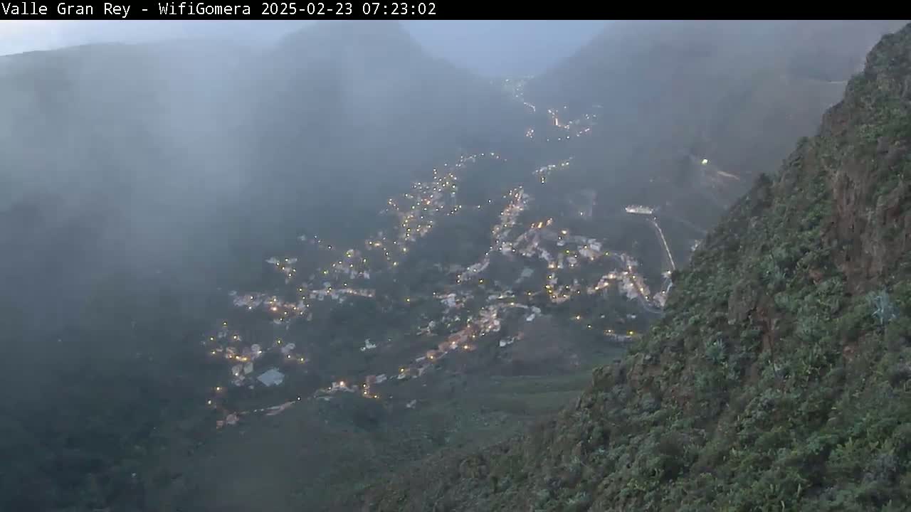 A foggy, nighttime view shows a town nestled in a valley between steep, dark green mountains, with many lights visible.