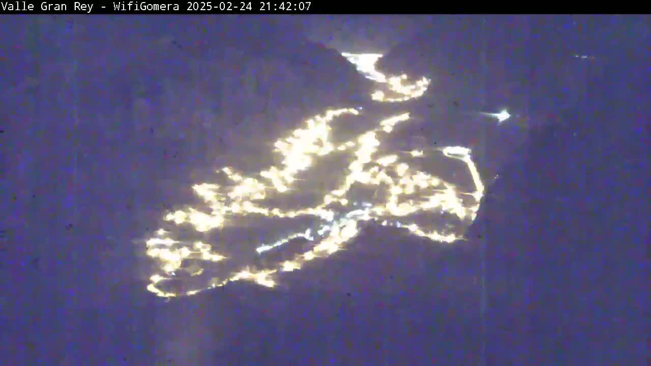 A nighttime, long-distance view shows the lights of a town or village situated on a dark, possibly mountainous landscape.