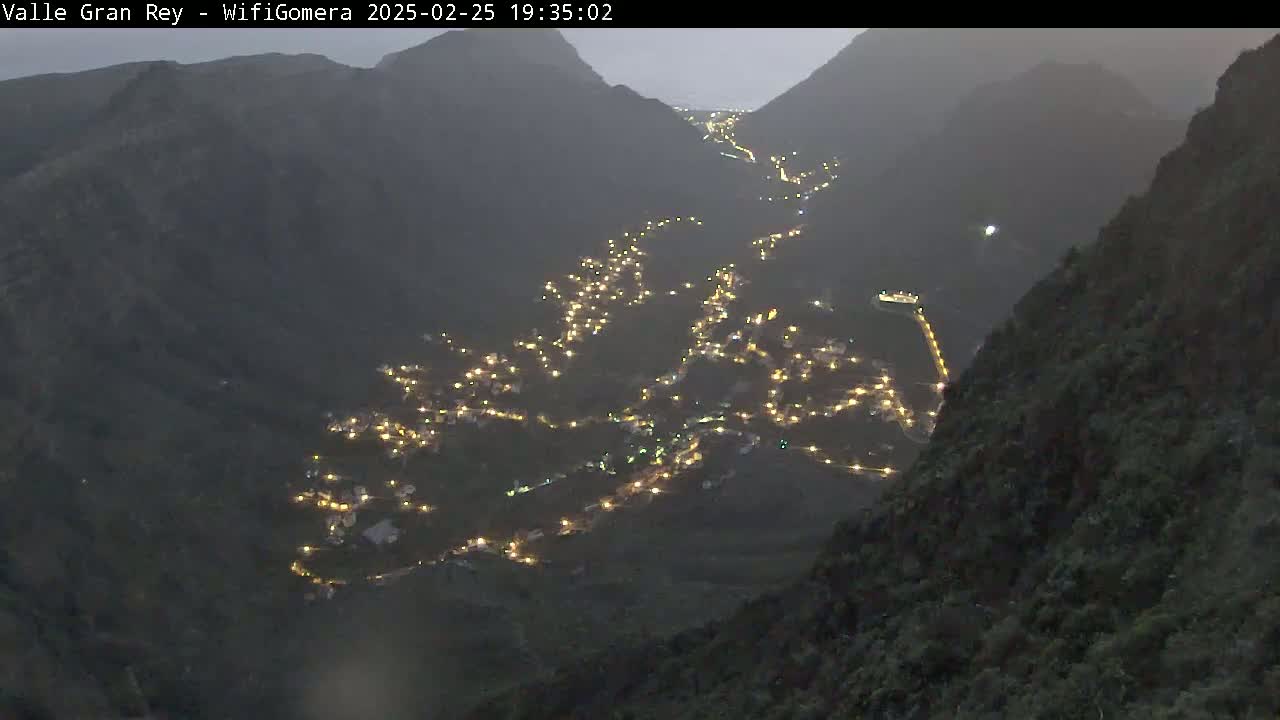 A valley nestled between dark mountains at night is illuminated by the lights of a town below.