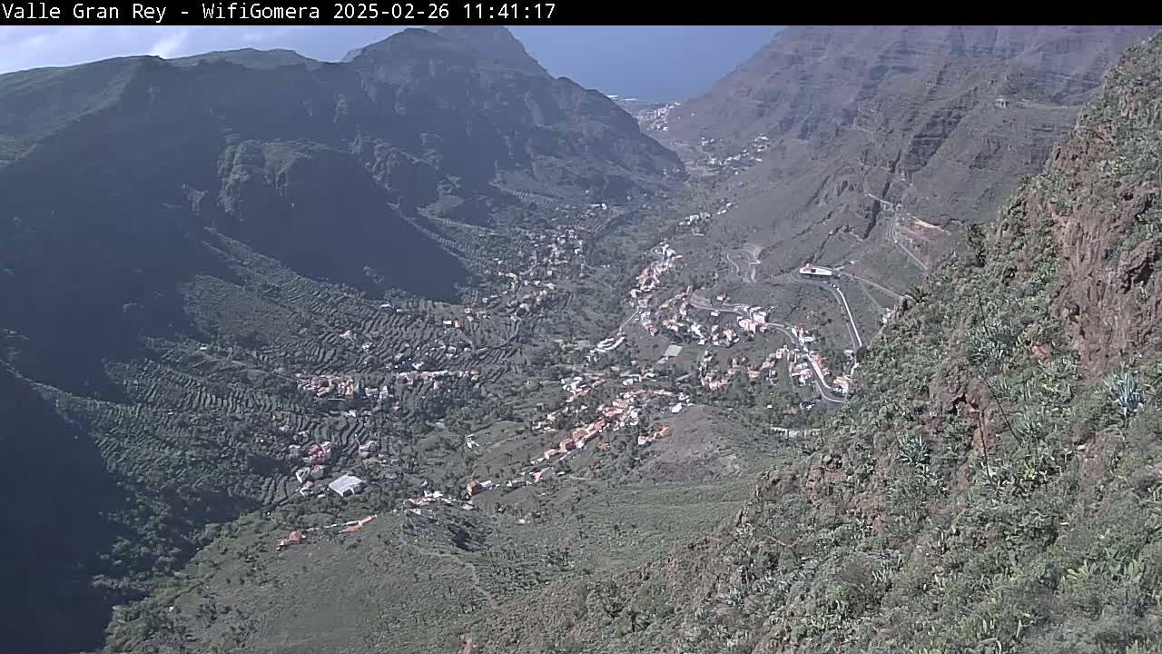 A high-angle view on a sunny day shows a town nestled in a mountainous valley, with terraced farmland surrounding it.