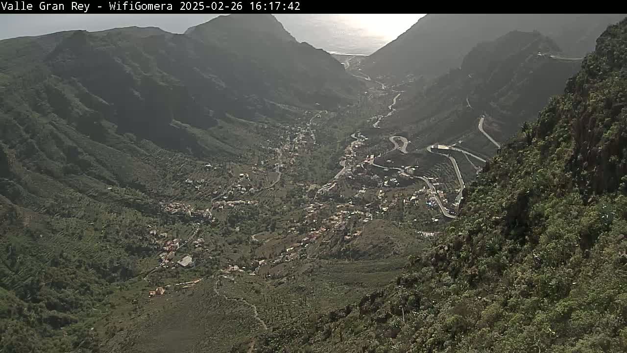 A hazy, high-angle view shows a village nestled in a verdant valley between steep, dark-colored mountains, with a winding road visible.