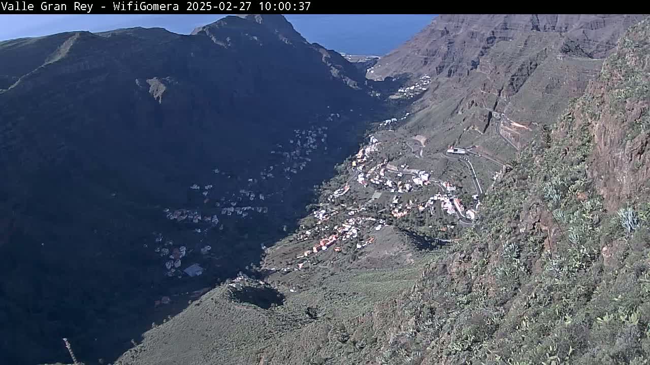 A sunny, high-angle view shows a village nestled in a deep valley between steep, rocky mountains, with a glimpse of the ocean in the distance.