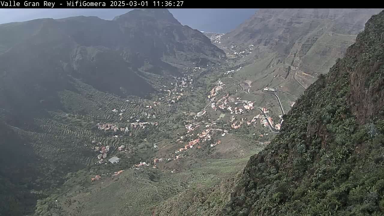 A hazy view shows a valley town nestled between steep, green mountainsides under a somewhat overcast sky.