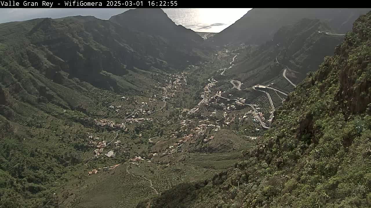 A high-angle view shows a village nestled in a deep valley between steep, green mountainsides, overlooking a distant ocean under sunny conditions.